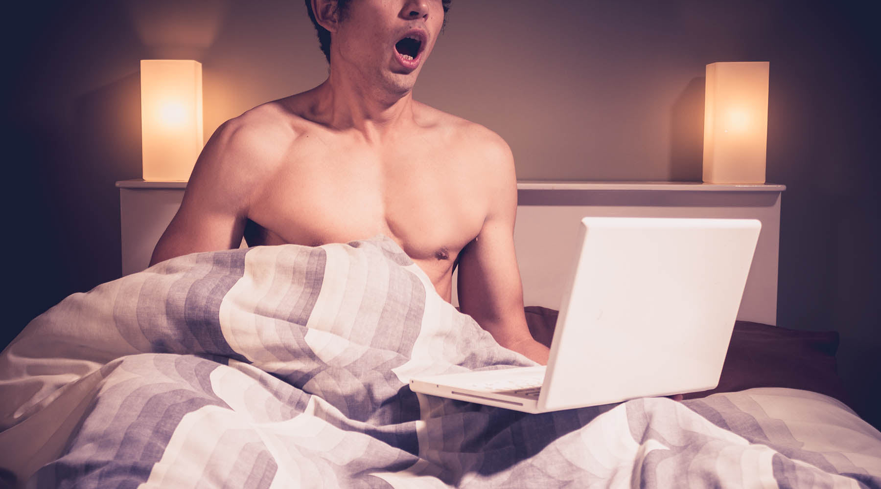 Shirtless man in bed with laptop that could benefit from masturbation training