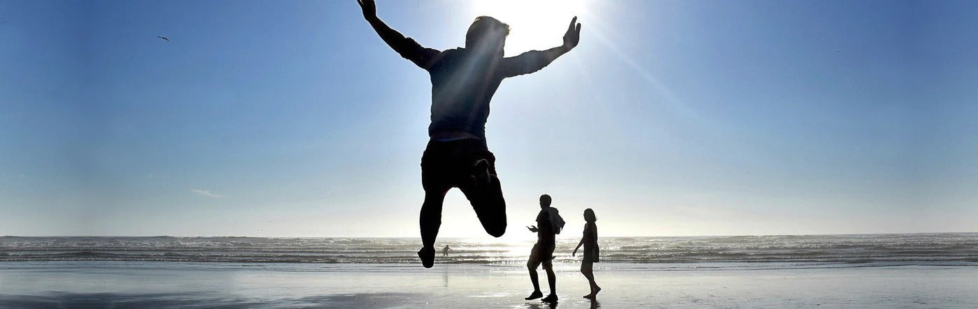 Man Jumping With Arms Out on Beach With Sun Behind Him With a Couple and the Sun Behind Him